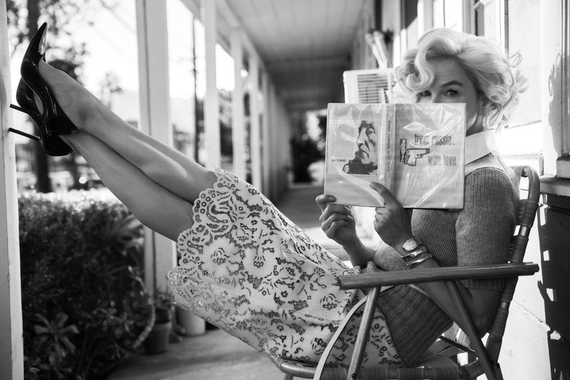 Blonde woman in heels and a dress sitting in a chair, holding a book in front of her face, on a porch in a black and white photograph.