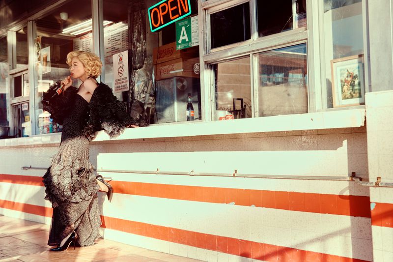 Woman in vintage dress and heels eats a lollipop outside a vintage diner with neon signs. Fashion editorial, sunny day, outdoor setting.
