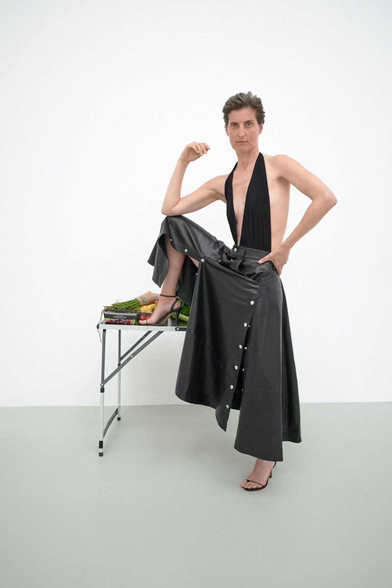 Adult female model poses in a black top and black skirt with a leg resting on a table covered in produce in a white studio.