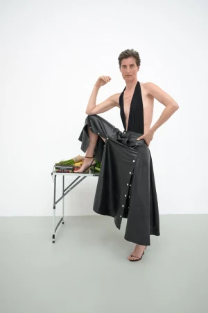 Adult female model poses in a black top and black skirt with a leg resting on a table covered in produce in a white studio.