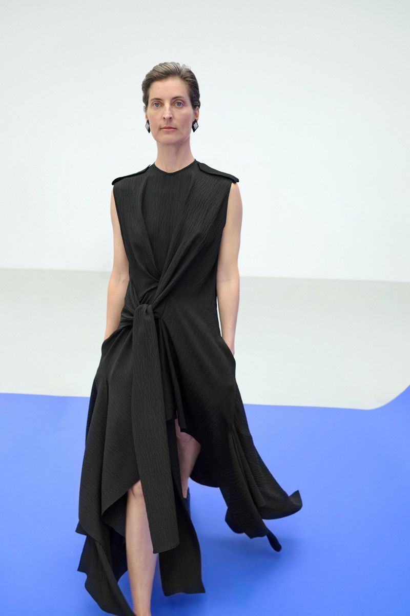 Woman in black dress poses in a fashion shot against a blue and white background, looking forward with a neutral expression.