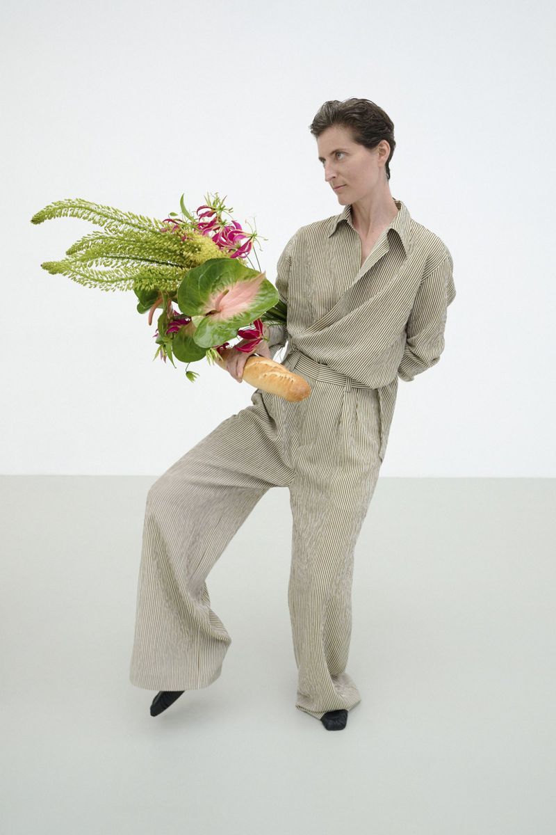 A person dressed in a tan-colored outfit holds a bouquet of flowers and a baguette in a white studio with a neutral expression.