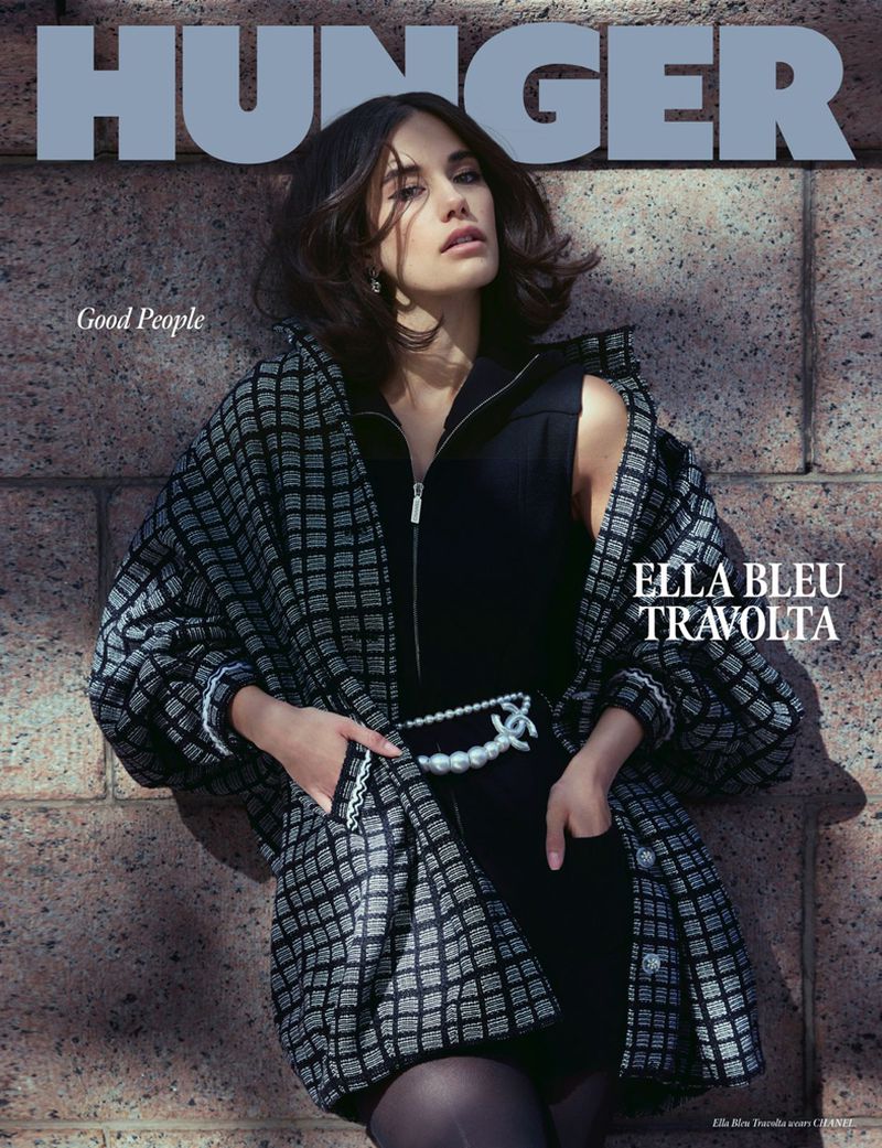 Ella Bleu Travolta graces the cover of Hunger Magazine in a portrait-style image. She's wearing a zip-up dress and patterned coat, leaning against a wall.