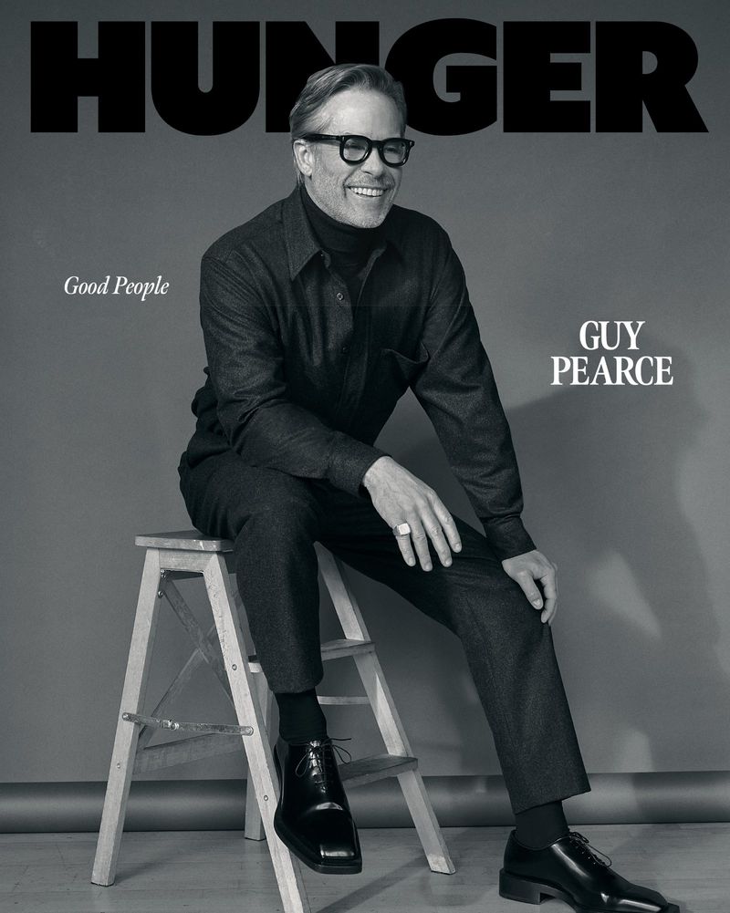 Black and white portrait of a man sitting on a step stool wearing a button-down shirt, turtleneck, dress pants, and dress shoes.