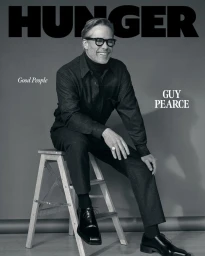 Actor Guy Pearce wearing glasses and dark monochromatic attire seated smiling on a wooden stepladder for a magazine cover.