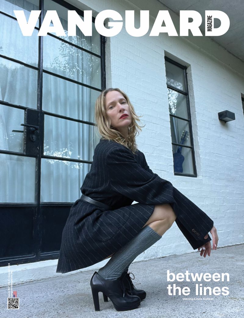 Magazine cover featuring a blonde woman in a pinstripe suit, kneeling outdoors against a white brick wall, styled with black platform heels.