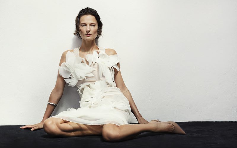 Adult woman sitting on a black surface against a white wall, wearing a light-colored dress with tulle and flower details and nude-colored shoes.