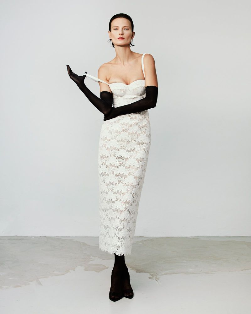 Model Tanya Ruban wearing a white lace midi dress and black opera gloves against a white studio background.