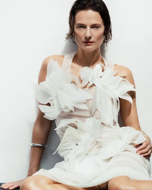 Elegant adult female model poses in a studio wearing a white dress with floral accents against a white backdrop.