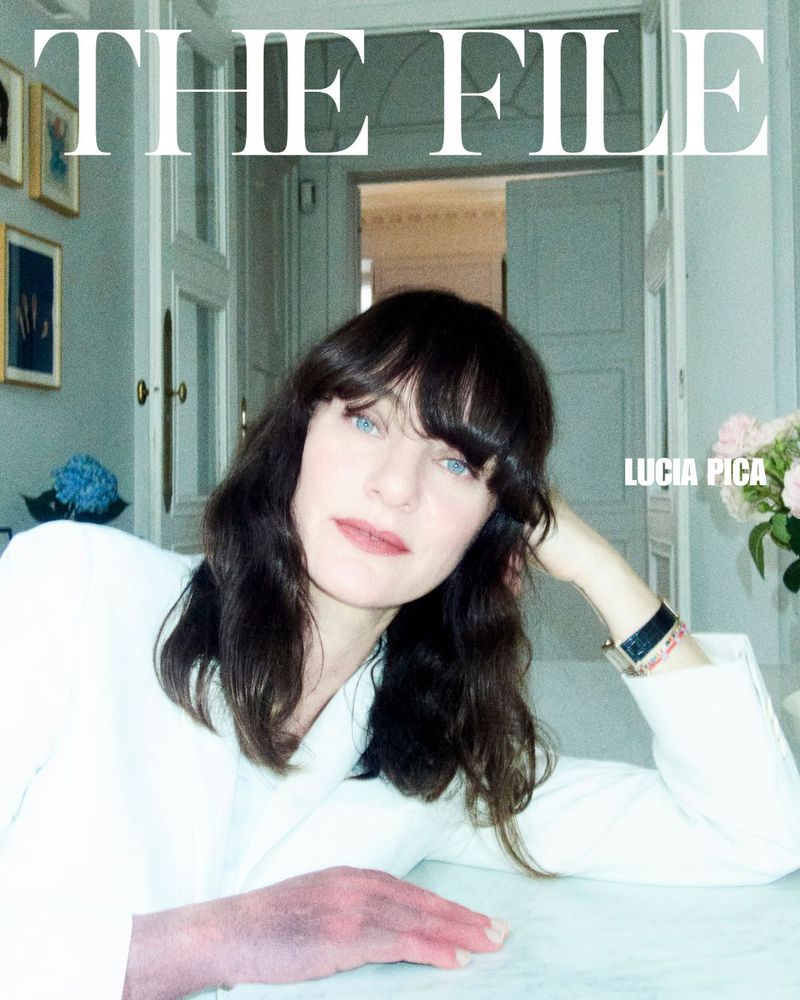 Close-up portrait of Lucia Pica with dark hair and light eyes, wearing a white jacket, seated at a table indoors near a window with flowers.
