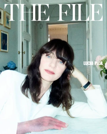 Close-up portrait of Lucia Pica with dark hair and light eyes, wearing a white jacket, seated at a table indoors near a window with flowers.