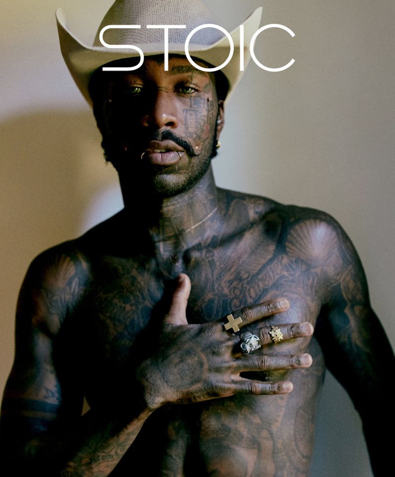 Portrait of a shirtless, tattooed adult male wearing a cowboy hat and rings, posing in a photo studio against a neutral backdrop.