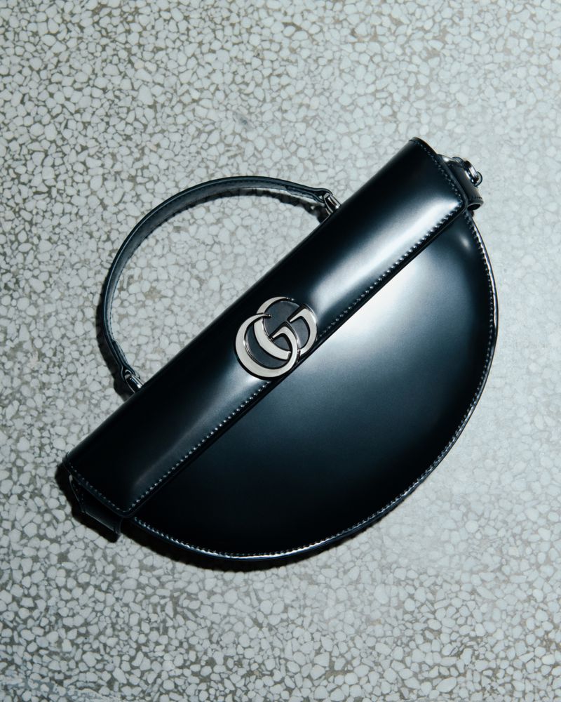 Gucci half-moon Jackie 1961 handbag in black patent leather with silver hardware, strap, and brand logo, photographed in a studio setting.
