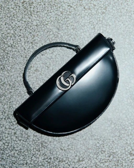 Gucci half-moon Jackie 1961 handbag in black patent leather with silver hardware, strap, and brand logo, photographed in a studio setting.