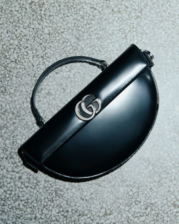 Gucci half-moon Jackie 1961 handbag in black patent leather with silver hardware, strap, and brand logo, photographed in a studio setting.