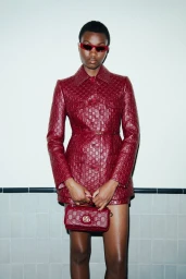 Model wearing a matching burgundy patterned leather jacket and holding a matching handbag against a tiled wall