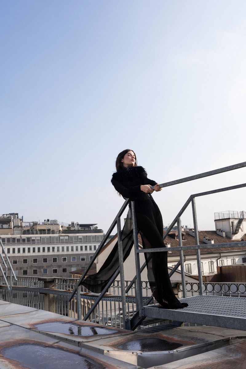 A woman in a black outfit stands on a metal platform overlooking a city skyline under a clear sky, fashion shoot, full shot, low angle.