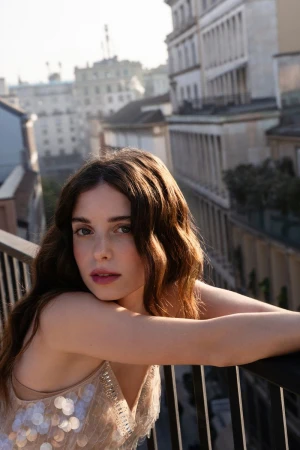 Brunette woman with shoulder-length wavy hair, poses on a balcony with cityscape background. Wearing a sequin-embellished top.