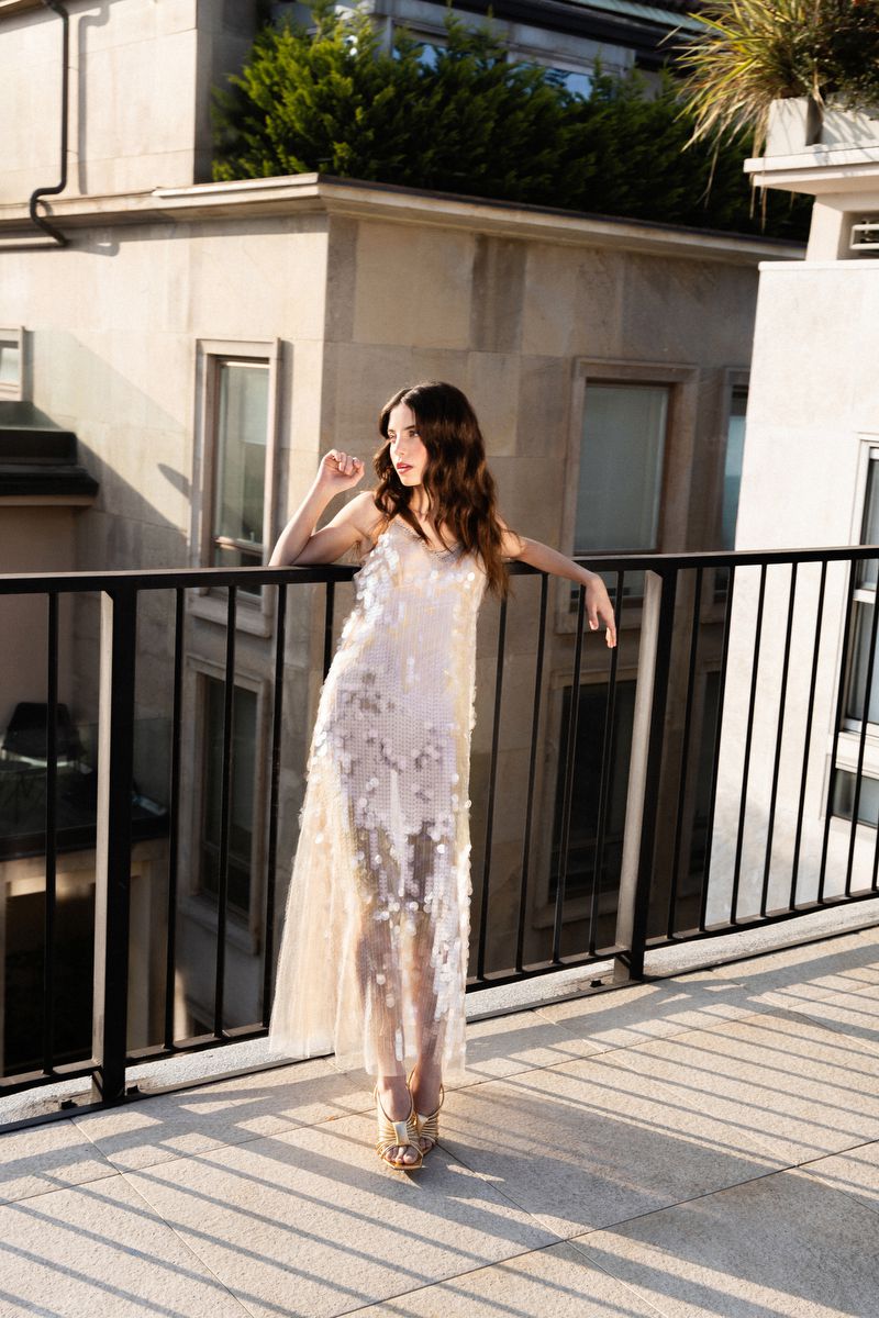Model wearing a sheer sequined floral slip dress posing against a black railing on a sunlit rooftop terrace.