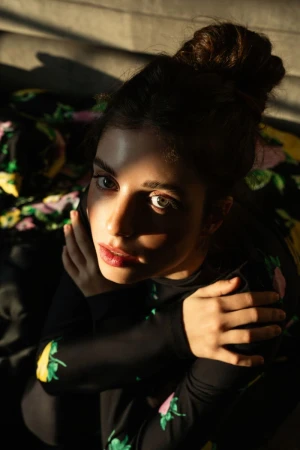 Close-up portrait of a woman with a high bun, wearing a black patterned top, illuminated by dramatic sunlight.