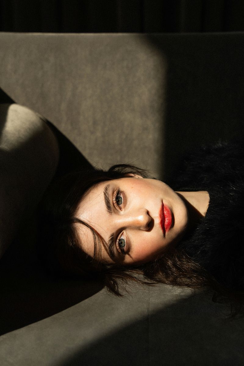 Close-up studio portrait of a young woman lying on gray couch under natural light, red lipstick, dark hair, gray background.