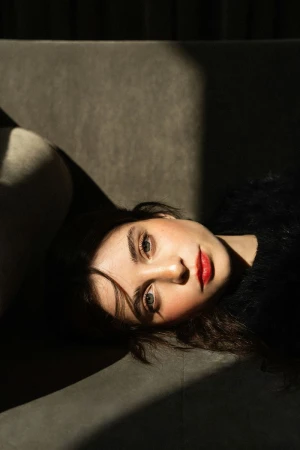 Close-up studio portrait of a young woman lying on gray couch under natural light, red lipstick, dark hair, gray background.
