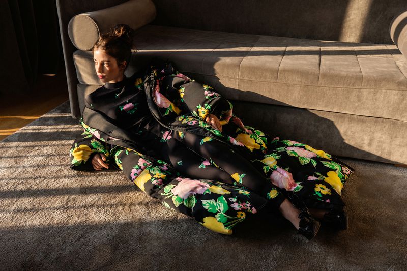 A woman in a floral outfit lies on a rug next to a grey couch in the sunlight, wearing black platform shoes and a matching floral outfit.