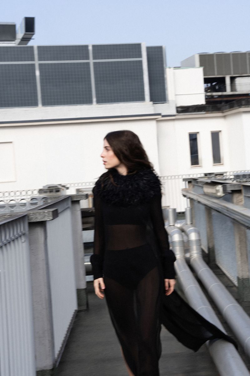 Woman in sheer black dress and feathery collar walks on rooftop, gazing left, against a backdrop of buildings and a light blue sky.