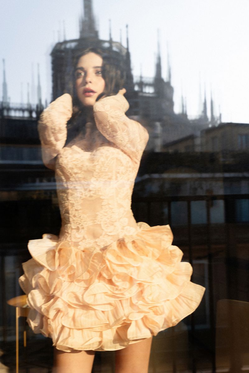 A young adult female poses in a peach-colored dress with a ruffled skirt, set against a blurred cityscape with gothic architecture.