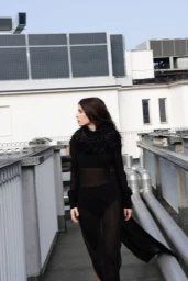 Woman wearing a sheer black dress with a feathered collar walking along a modern building rooftop walkway.