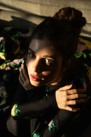 Close-up portrait of a woman with a high bun, wearing a black patterned top, illuminated by dramatic sunlight.