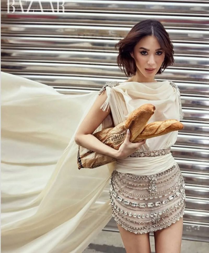 Woman in a dress poses holding baguettes against a metal background, promoting Bazaar magazine with a fashion editorial style.