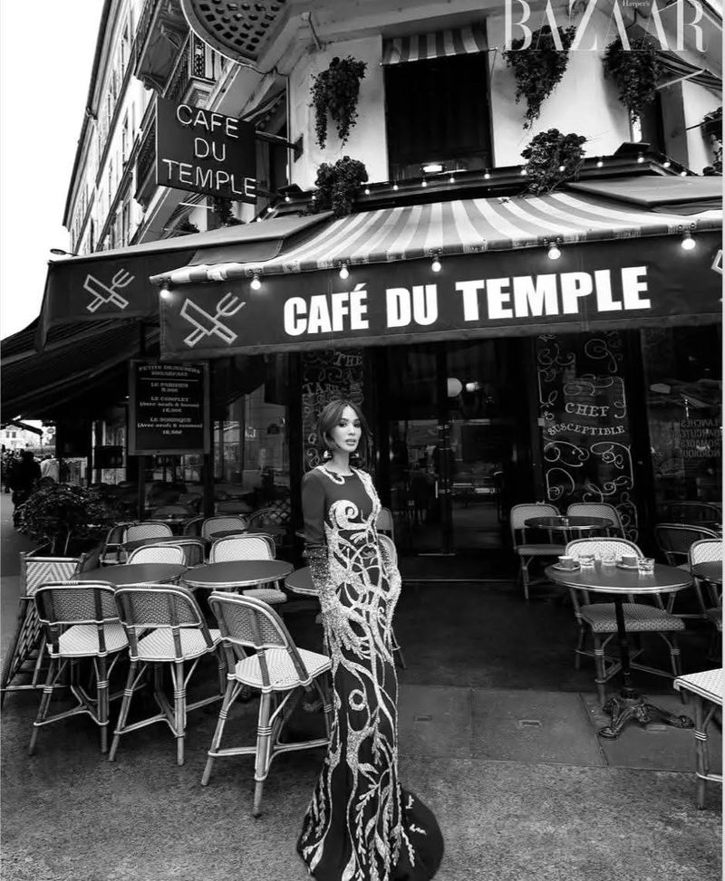 Adult female in a long evening gown poses in front of an outdoor cafe with tables and chairs in black and white with BAZAAR logo.