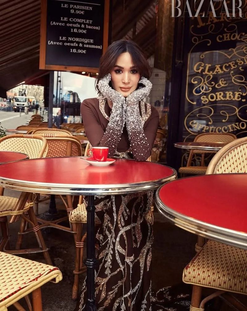 Woman in elegant brown gown with silver accents poses at outdoor cafe with red table, cup of coffee, Bazaar magazine in background.