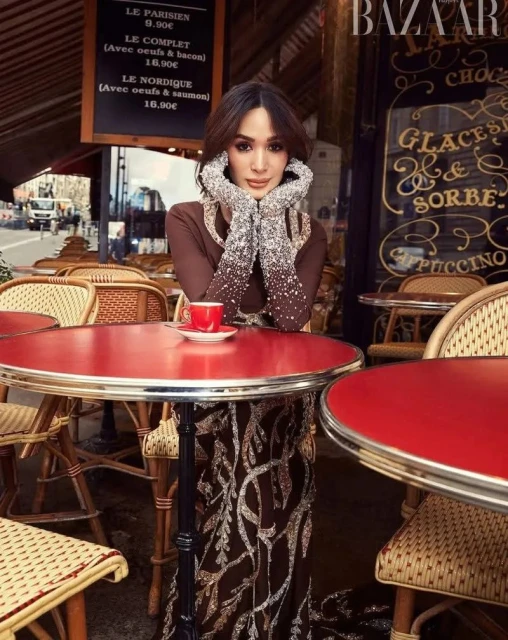 Woman in elegant brown gown with silver accents poses at outdoor cafe with red table, cup of coffee, Bazaar magazine in background.