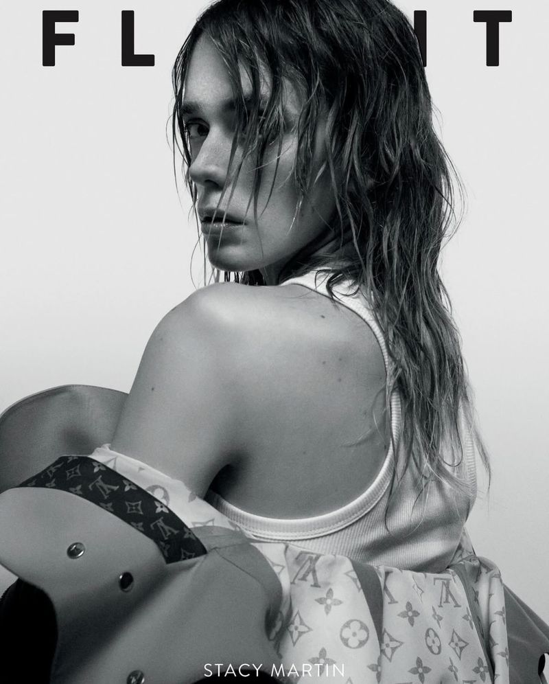 Black and white magazine cover shot of Stacy Martin looking back over her shoulder with wet hair and a jacket with logo print.