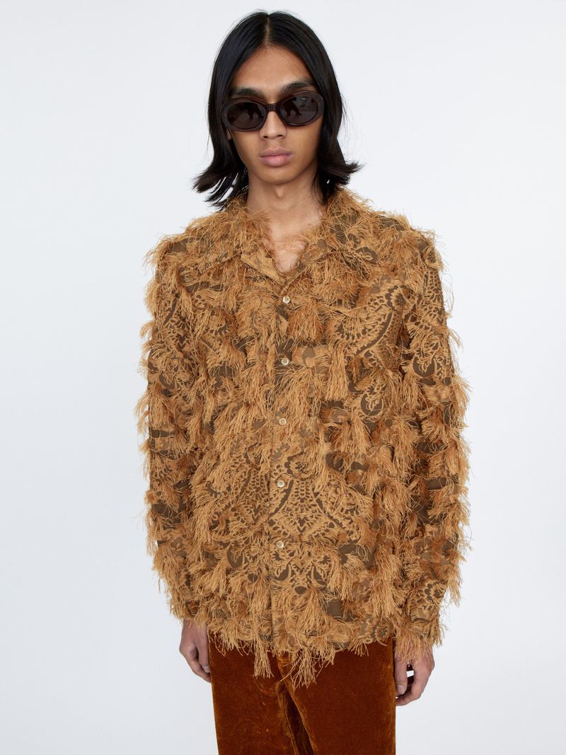 Man wearing fringed brown button-down shirt and sunglasses in a studio. Fashion shot with medium framing, solid white background.