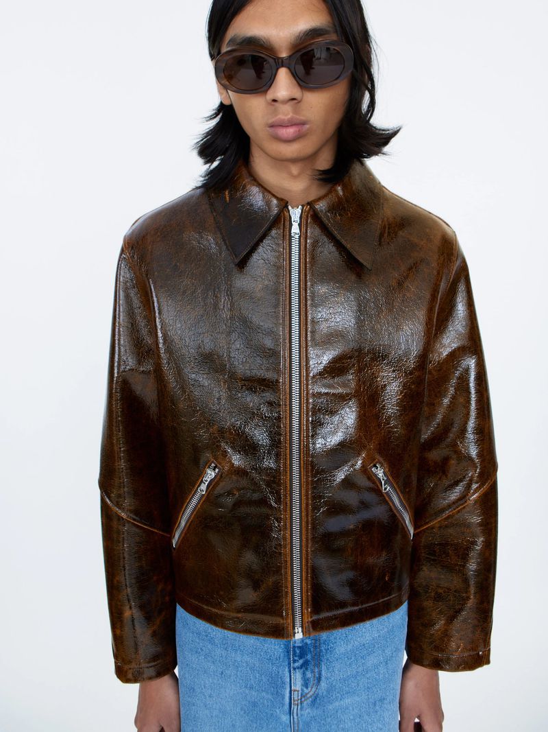 Model wearing dark oval sunglasses, a glossy brown leather jacket, and blue jeans against a plain white background.