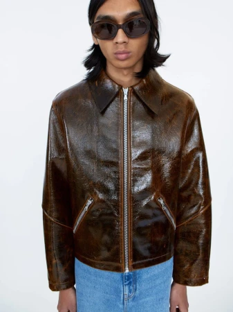 Model wearing dark oval sunglasses, a glossy brown leather jacket, and blue jeans against a plain white background.