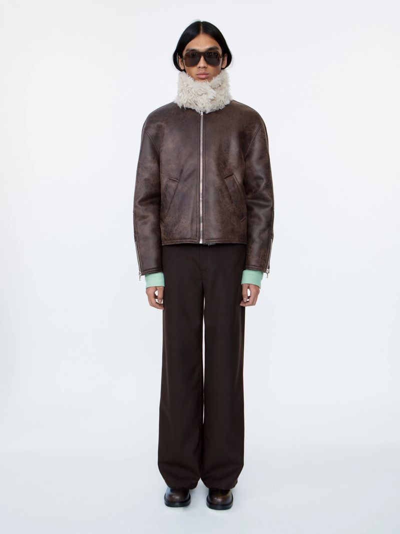 Full-length studio shot of adult male model wearing sunglasses, brown shearling coat, brown pants against a white backdrop.