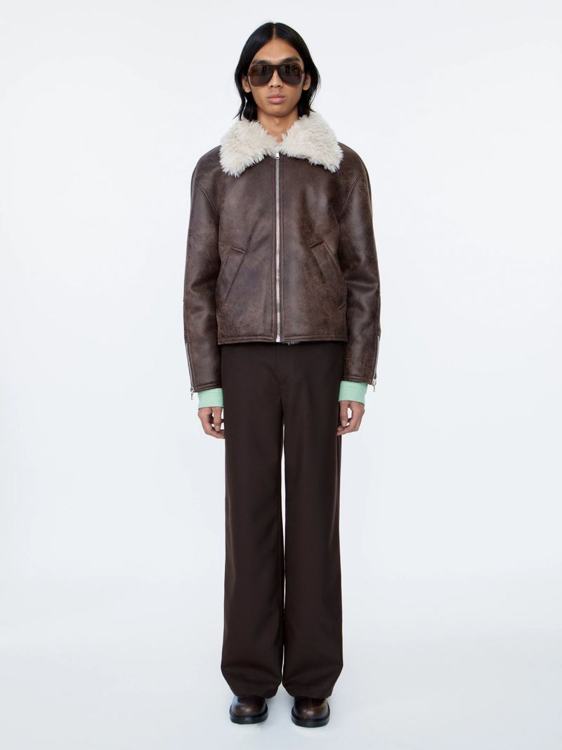 An adult male fashion model stands against a white backdrop, wearing a brown leather shearling bomber jacket and brown pants.