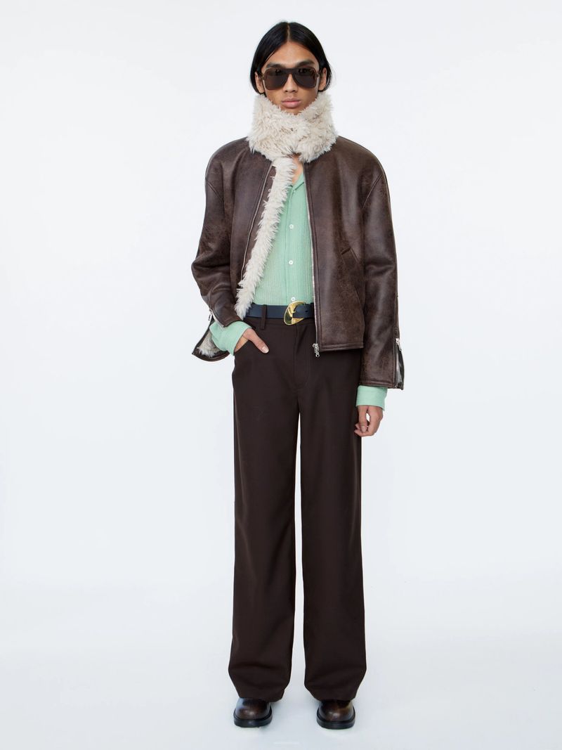 Full-length shot of a person in brown jacket and trousers, accessorized with sunglasses and scarf, against a white backdrop.