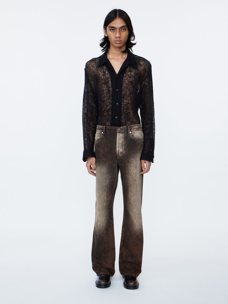 Adult male stands against a white background wearing a lace shirt, brown jeans, and dress shoes in a full-length portrait.