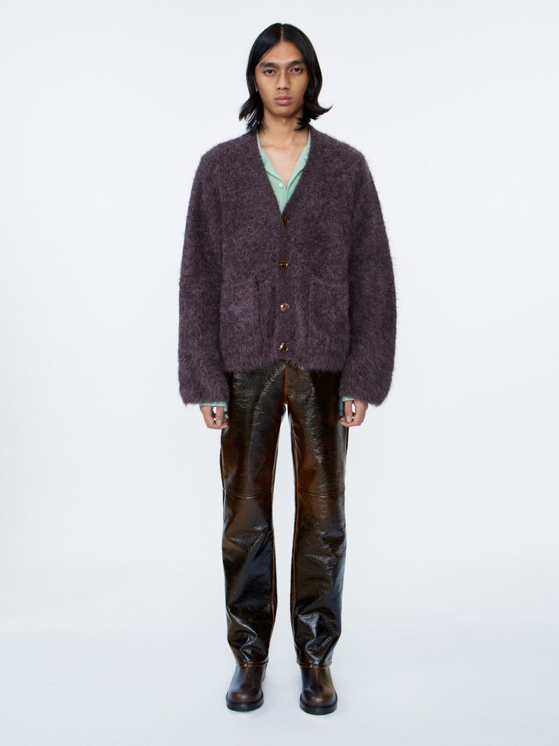 Full-length studio shot of a male model wearing a brown cardigan sweater, shiny brown pants, and shoes against a white background.