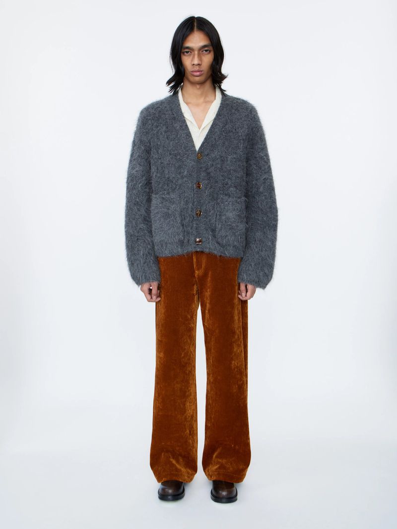 Studio shot of androgynous person wearing a fuzzy grey cardigan, off-white collared shirt, and velvet brown trousers, standing against a white backdrop.