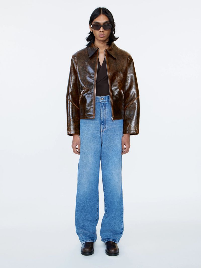 A full shot of a person wearing a brown jacket, blue jeans, sunglasses, and black shoes, standing against a white background in a studio.