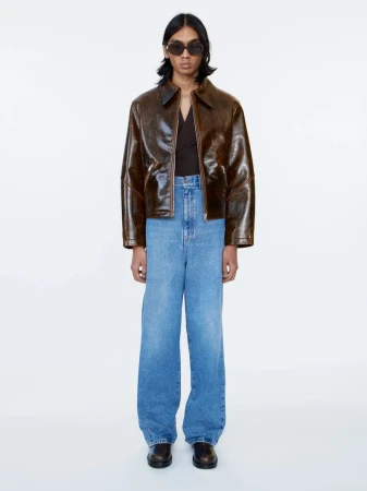 A full shot of a person wearing a brown jacket, blue jeans, sunglasses, and black shoes, standing against a white background in a studio.