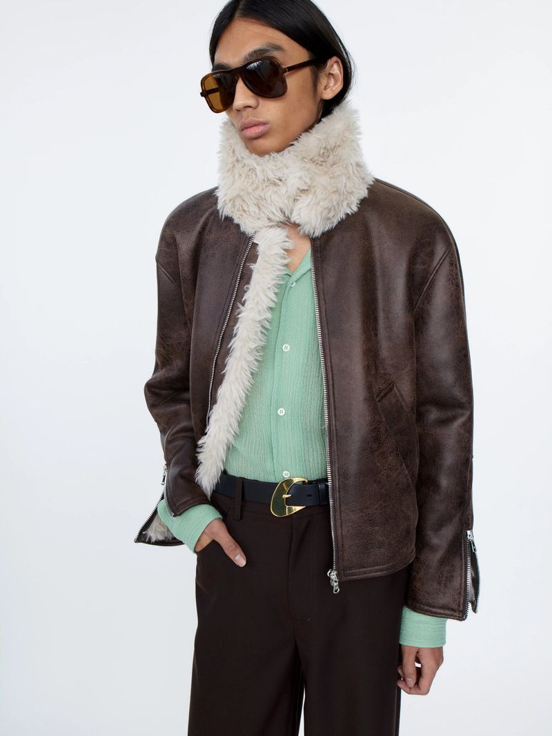 Adult male fashion model wears a brown leather jacket with fur collar, green shirt, brown pants, sunglasses against a white background.
