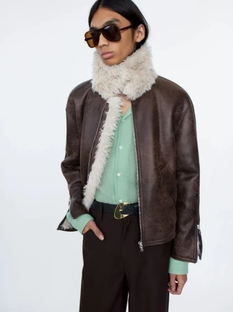 Adult male fashion model wears a brown leather jacket with fur collar, green shirt, brown pants, sunglasses against a white background.