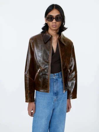 Model wearing a glossy brown leather jacket and blue jeans against a white backdrop